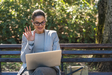 Professional woman sitting on park bench, focused on laptop work having video call with colleague,...