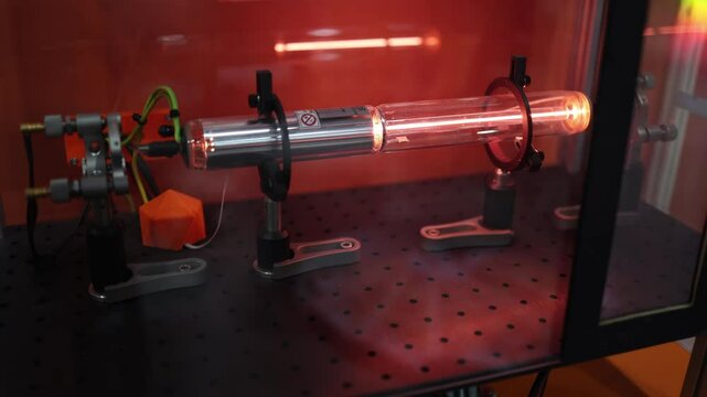 High-power laser tube glowing red in a precision lab setup with mounted components and safety casing shows cutting-edge technology for industrial laser systems or scientific research environments