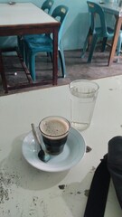 A cup of black coffee is on a small plate with a spoon, next to a glass of water, in a simple room with light blue walls.