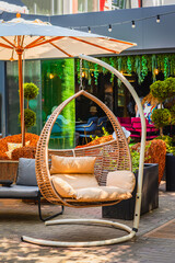 Wicker hanging chair with beige cushions in stylish outdoor cafe surrounded by greenery and cozy decor. Concept of outdoor comfort, relaxation space, modern lifestyle