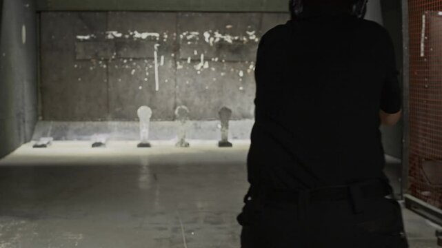 Back view tracking shot of man wearing ear protectors and goggles practicing movements with rifle and shooting at indoor range