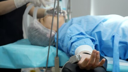A female patient lies on a surgical operating table in a clinic with a pulse monitor on her finger, a doctor puts on a ventilator mask. - Powered by Adobe