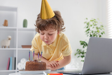 Birthday celebration. Little boy blowing candles on cake while having video call via laptop at...