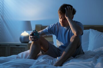 Frustrated man looking at alarm clock on bed at night. Insomnia problem