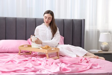 Woman pouring coffee from moka pot into cup on bed with pink silk linens indoors