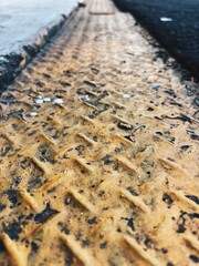The image shows a close-up, low-angle view of a weathered, textured yellow metal surface with a diamond plate pattern, bordering an asphalt road.
