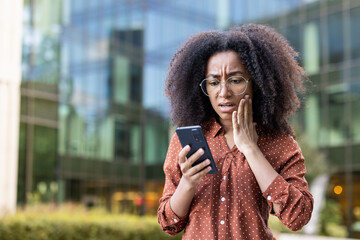 An anxious businesswoman in glasses reacts with concern while looking at her phone in an urban...