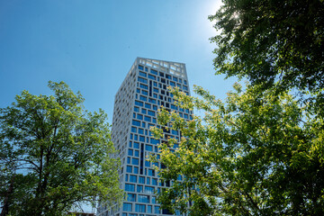 Fototapeta premium A tall building with geometric window patterns, surrounded by green trees, against a clear blue sky. Modern office building