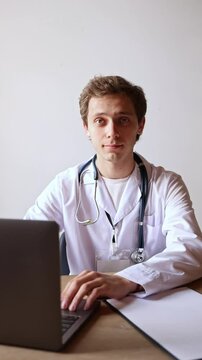 Male doctor in white coat sits at desk, typing on laptop with concentrated expression, working with digital records in clinic. Concept of online consultation, diagnostics, workflow.