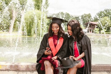 Two female graduates using smartphone near fountain
