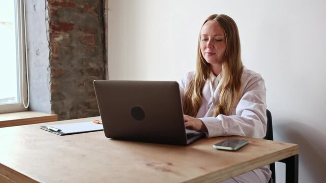 Focused woman in white coat types on keyboard, completing digital forms or reports, working efficiently in private office space. Concept of e-medicine, professional care, patient records.