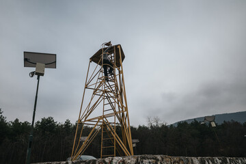 An observation tower structure situated outdoors, under an overcast sky, with a forest backdrop and...
