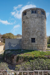 An ancient stone tower, possibly a windmill or defensive structure, standing tall under a bright blue sky with fluffy clouds, surrounded by Mediterranean vegetation.