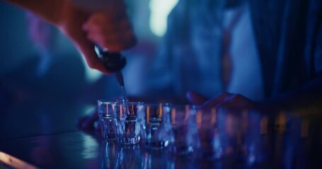 Close Up of a Bartender Pouring Clear Liquor Into Shot Glasses Lined Up on the Bar Table. Hand Moves Comfortably Between Glasses Without Spilling the Spirit Under Colorful Nightclub Lights - Powered by Adobe