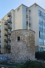 An ancient stone tower stands prominently in front of a modern multi-story hotel building, showcasing the architectural blend of Rhodes, under a clear sky.