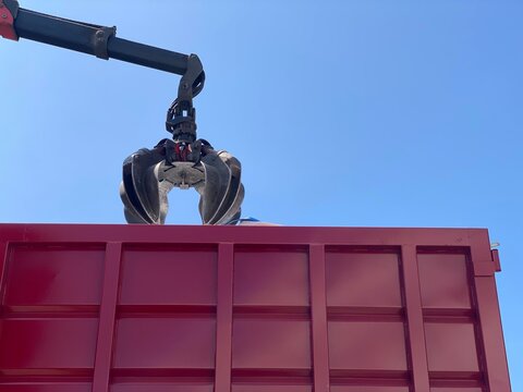 Hydraulic industrial claw grabs metal scrap over a red container against clear blue sky. Heavy machinery in use for waste management, recycling or construction work.