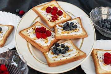 Flat lay of toasted white bread topped with labneh, fresh raspberries, blueberries, maple syrup, and flax seeds. Sweet breakfast or brunch idea with natural light and food styling