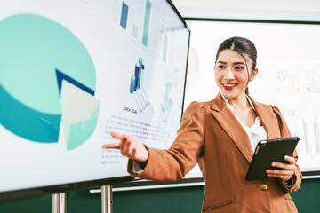 Confident businesswoman in a brown suit presenting financial charts on a large screen with a digital tablet in hand, showcasing analytic, teamwork, and strategic corporate planning in the office.
