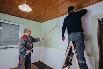 Two men collaborating on a home improvement project, applying paint to a room's walls with rollers....