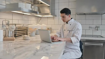 A young Asian man in a chef's uniform works on a laptop in a modern kitchen. The space features stainless steel appliances and a marble countertop.