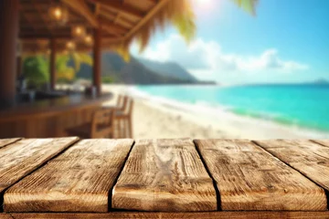 Fotobehang Zomer Empty wooden table with open space for product placement or branding. In the background blurred summer scene with sandy coastline and beach cafe and the blue sea under sunny sky.  © magdal3na