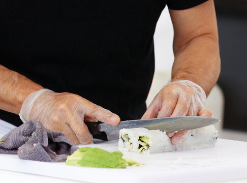 Close-up view of a sushi chef slicing a roll on a white cutting board, showcasing professional culinary preparation while wearing protective gloves to ensure hygiene in meal preparation.