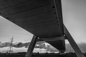 Low-angle black and white view beneath the concrete structure of the Flintshire Bridge, with distant pylons silhouetted against a cloudy sky