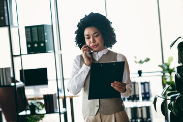 Confident businesswoman reviewing documents while managing a phone call in a modern and...
