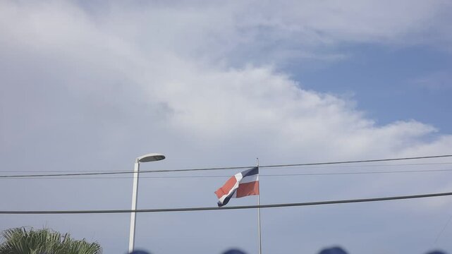 dominican republic flag waving under a cloudy blue sky in an urban setting, with visible street lights and power lines crossing the frame, canon eos R c-log color 8bits