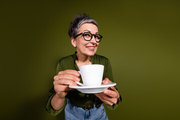 Charming mature woman wearing stylish glasses holding a cup and saucer with a cheerful smile against a green background