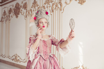 Young woman in a pink baroque costume admires her reflection with an elegant hairstyle and makeup