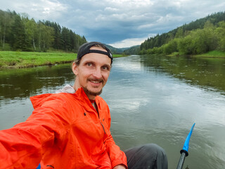 A man in an orange jacket is taking a selfie in a boat on a river