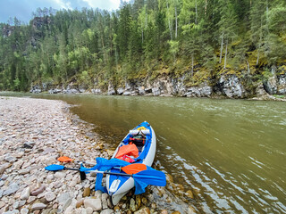 A bright and colorful kayak is sitting quietly on the sandy shore of a flowing river, waiting to be taken out for an adventure on the water. The scenery is serene and beautiful
