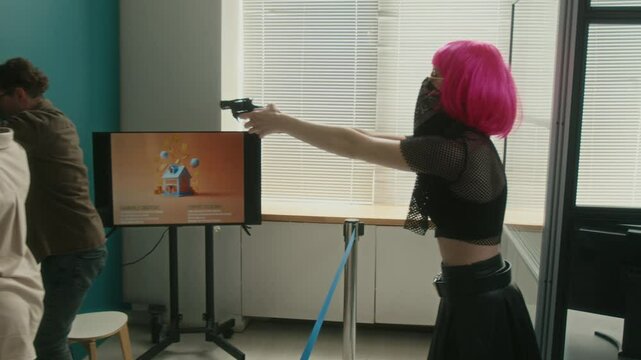 Female criminal masked with bandana and pink wig pointing handgun at bank customers telling hostages to turn away with arms raised while male accomplice demanding money from teller