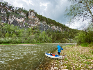 A man is standing on the peaceful shore of a river, right next to a sturdy raft that is moored nearby, enjoying the tranquil beauty of the natural environment and watercourse