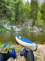 A person is comfortably seated next to a vibrant blue and white kayak that is resting on the sandy shore of a tranquil river, enjoying the peaceful beauty of nature all around them