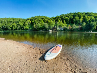A colorful paddle board is currently resting on the warm sandy shore of a tranquil lake, waiting for someone to take it out onto the peaceful water surface for some fun and adventure