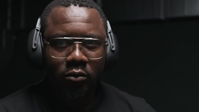 Zoom in portrait of young black man in glasses and earmuffs confidently looking at camera during training with rifle in shooting club