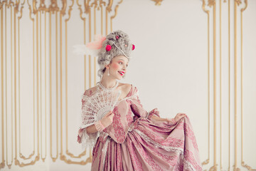 Elegant young woman in pink period gown holding lace fan inside luxurious historical palace room with ornate decor
