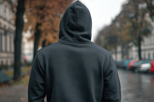A man in a black hooded sweatshirt walks away on a rainy street, the back view shows the hoodie and surroundings including trees and parked cars. - Powered by Adobe