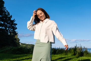 A bob-haired girl in a white shirt and green skirt adjusts her hair in the Norwegian outdoors. A gentle breeze, calm, and beauty in a simple gesture.