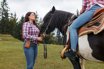 A woman holds the reins while another sits on a black and white painted horse in a green field....