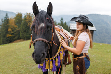 Obraz premium A young woman in western wear stands with her dark horse in a grassy pasture. Mountains and trees are in the background on a cloudy day in Europe.