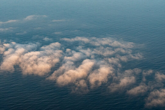 Aerial view showcasing soft, fluffy clouds tinged with warm, golden hues as they hover above the vast, deep blue expanse of the Pacific Ocean. The sunlight creates a gentle contrast