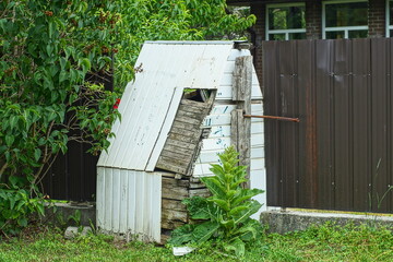 one wooden with hole closed with iron curved handle with white broken siding public deep abandoned well stands on ground near brown fence and tree with green leaves