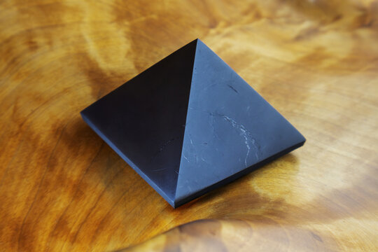 Black shungite pyramid sits on a wooden surface. The pyramid is made of a dark material and has a sharp point at the top. The wooden surface is brown and has a grainy texture