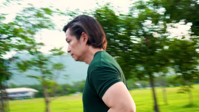Sports asian man running on road for outdoor workout with warm light of sunset on freshy green of nature  background in rainy season. Jogging in countryside. Healthy lifestyle. 