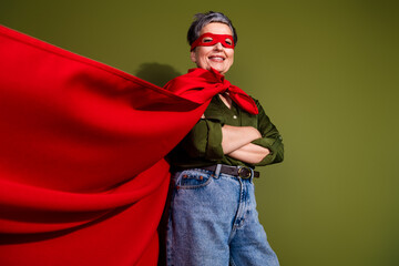 Confident senior woman in red superhero cape smiling against green wall in casual attire, showing...