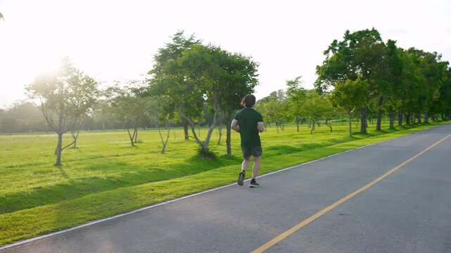 Sports asian man running on road for outdoor workout with warm light of sunset on freshy green of nature  background in rainy season. Jogging in countryside. Healthy lifestyle. 