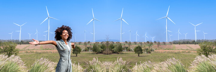 African american cheerful young woman relaxing with wind turbine farm background, Wind turbines are...